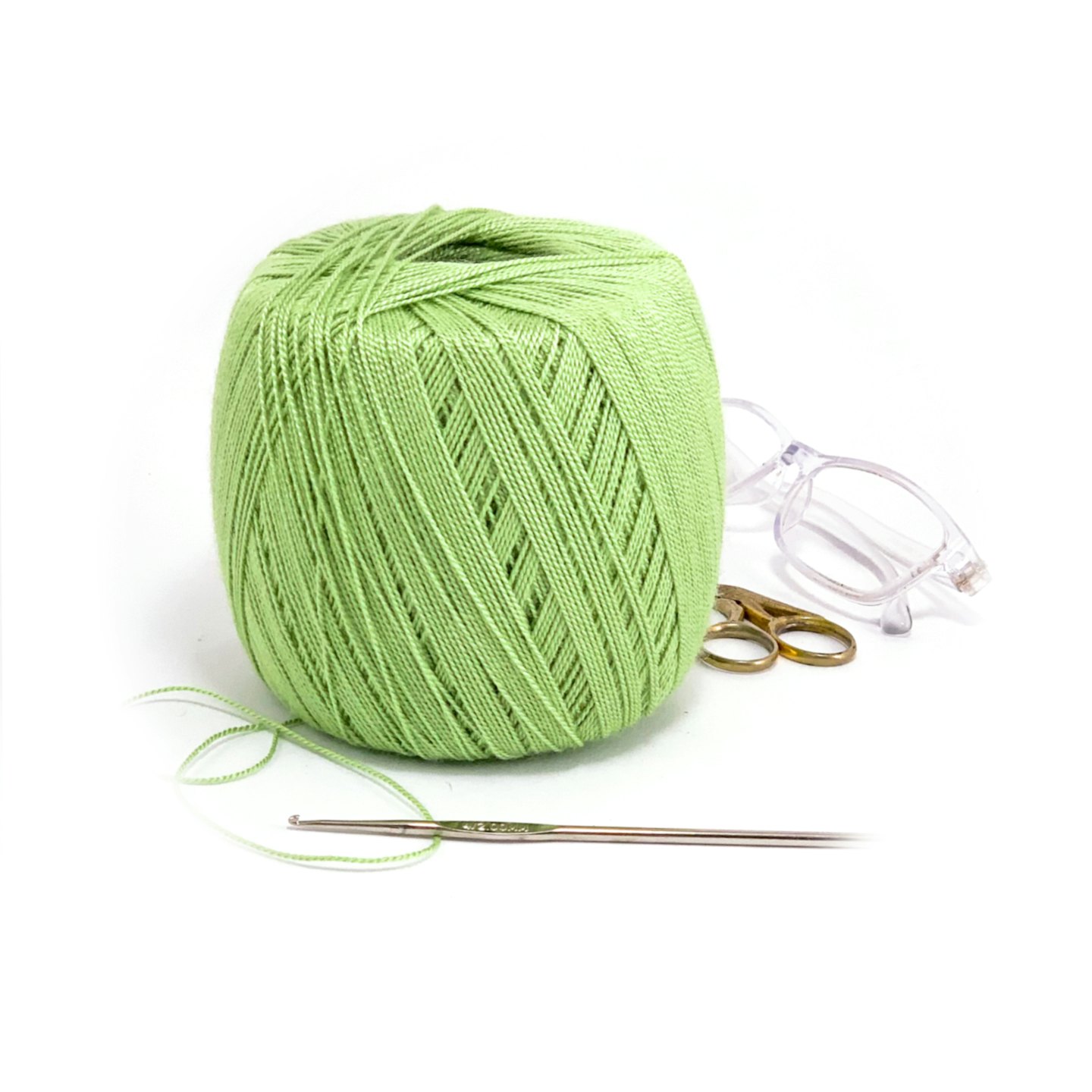 Green yarn, crochet hook, scissors, and glasses on white surface.